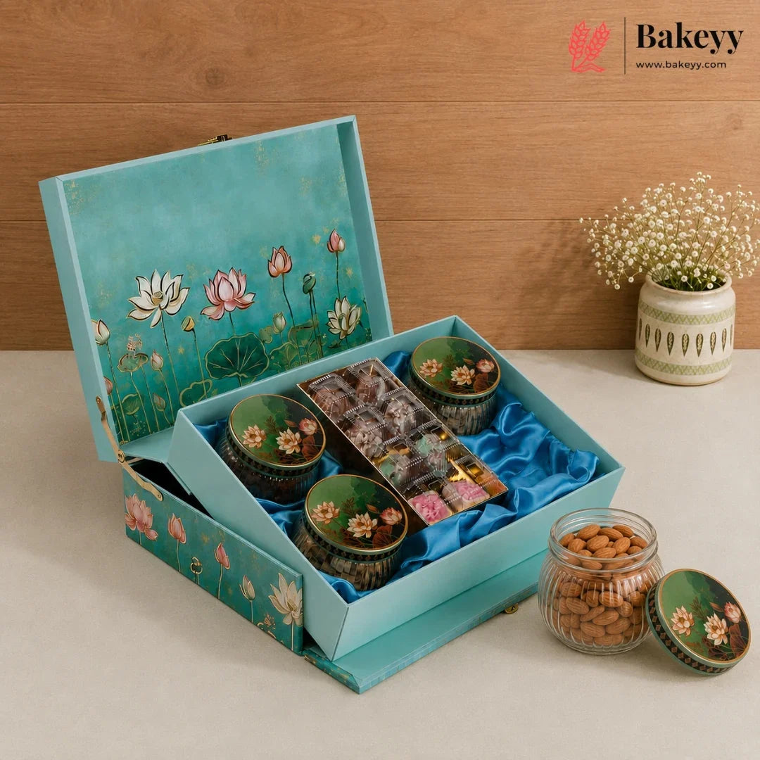 A teal and pink lotus-themed gift box from Bakeyy, filled with assorted chocolates and sweets, displayed on a beige surface against a wooden background with a small potted plant.