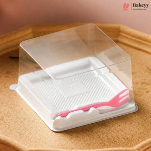 A clear plastic square food storage container with a white textured base and a small pink fork resting inside, shown on a tan surface. The Bakeyy logo appears in the top right corner.