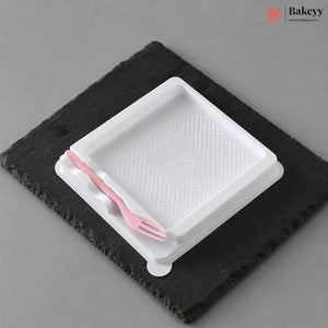 A white plastic tray with a textured interior and a small pink plastic fork resting inside, placed on a dark charcoal slate coaster against a neutral gray background.