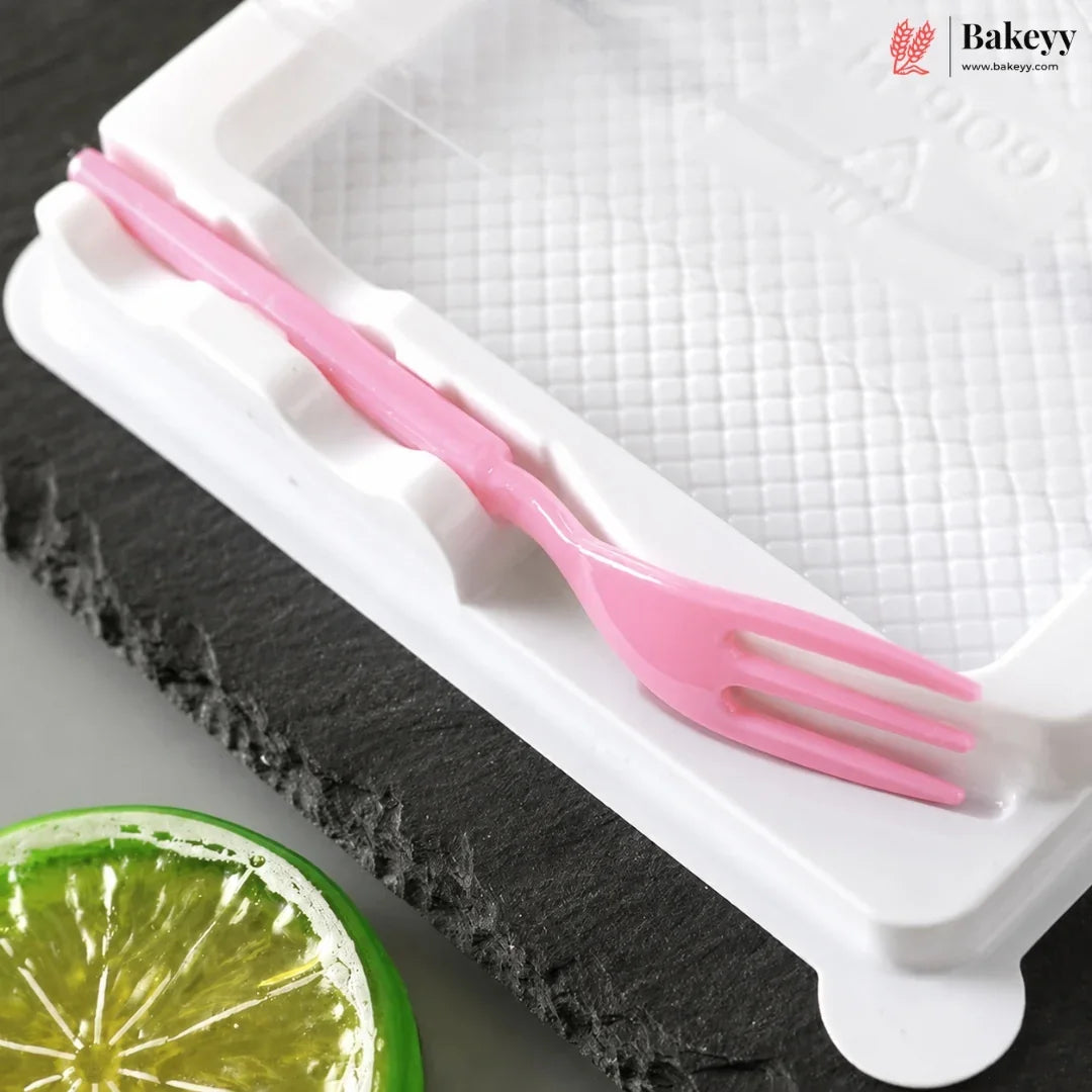 A pink plastic fork-shaped utensil resting on a white textured baking mold tray, with a dark slate board and a lime slice in the background. The Bakeyy logo is visible in the top right corner.