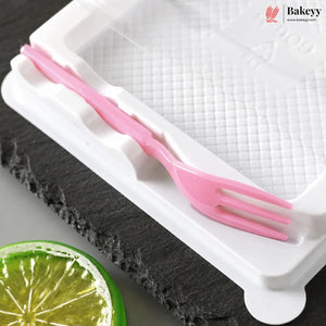 A pink plastic fork-shaped utensil resting on a white textured baking mold tray, with a dark slate board and a lime slice in the background. The Bakeyy logo is visible in the top right corner.