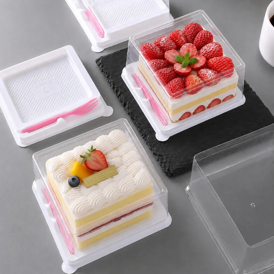 Clear plastic square cake boxes with white lids, shown with two decorated layered cakes on a gray tabletop. One box contains a strawberry-topped cake and the other a cream-colored cake with fruit garnish.