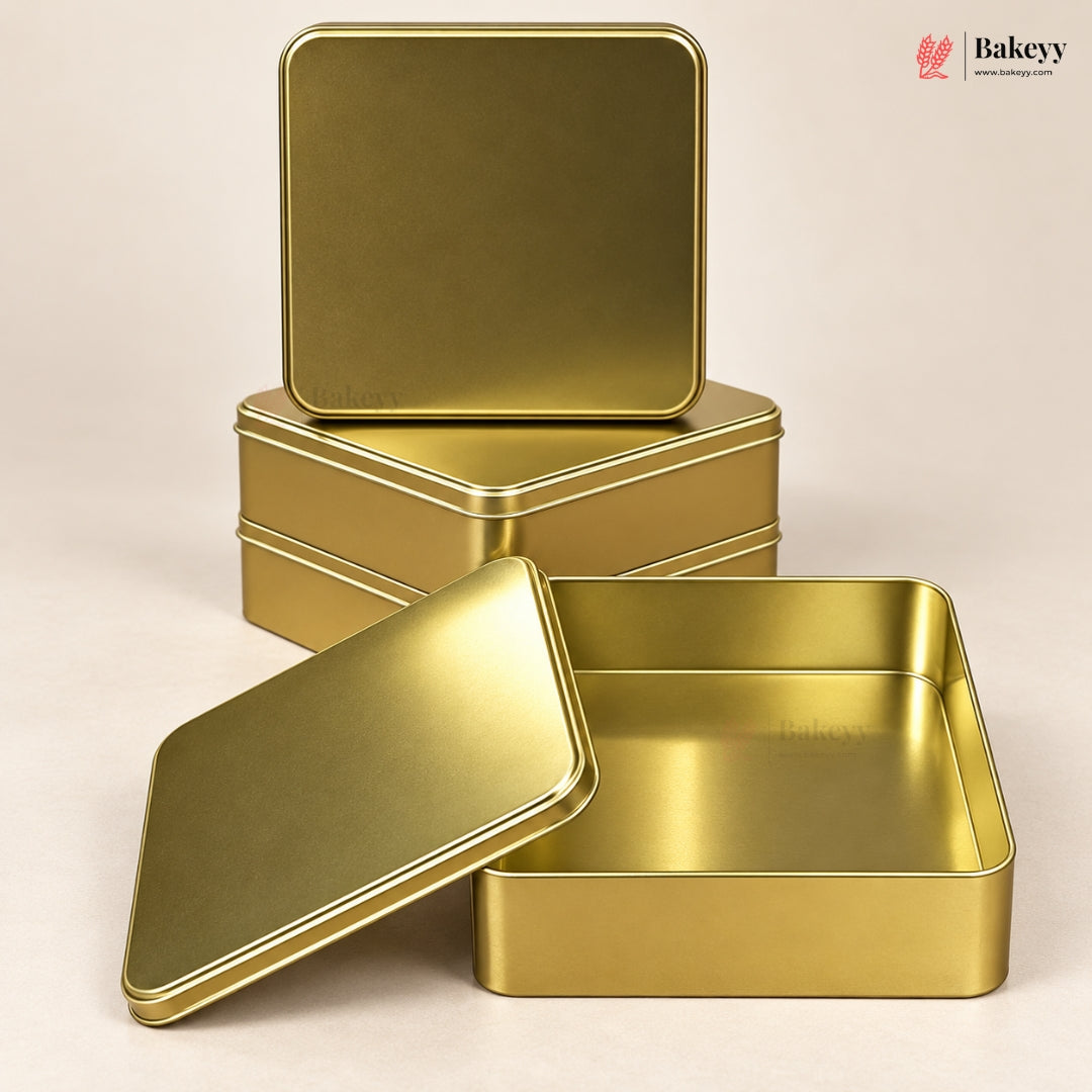 Three gold-colored metal tin boxes arranged on a light beige background, with one open box in the foreground and two stacked closed boxes behind it. A small Bakeyy logo appears in the top right corner.