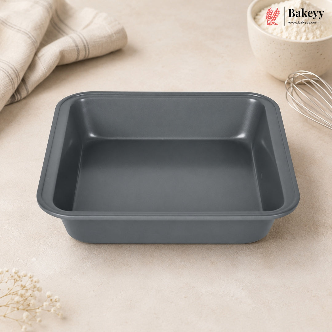 A dark gray rectangular non-stick baking pan with raised edges, shown on a light beige countertop with a folded cloth, a bowl of flour, and a whisk nearby. The Bakey logo appears in the top right corner.
