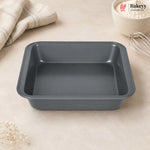 A dark gray rectangular non-stick baking pan with raised edges, shown on a light beige countertop with a folded cloth, a bowl of flour, and a whisk nearby. The Bakey logo appears in the top right corner.