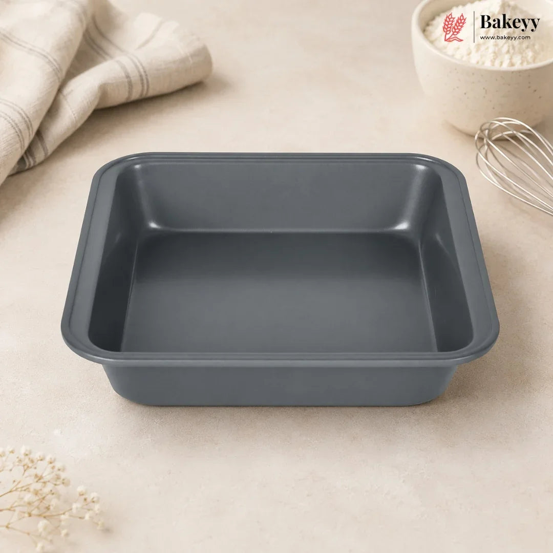 A dark gray rectangular non-stick baking pan with raised edges, shown on a light beige countertop with a folded cloth, a bowl of flour, and a whisk nearby. The Bakey logo appears in the top right corner.