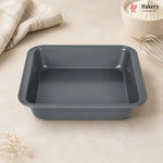 A dark gray rectangular non-stick baking pan with raised edges, shown on a light beige countertop with a folded cloth, a bowl of flour, and a whisk nearby. The Bakey logo appears in the top right corner.
