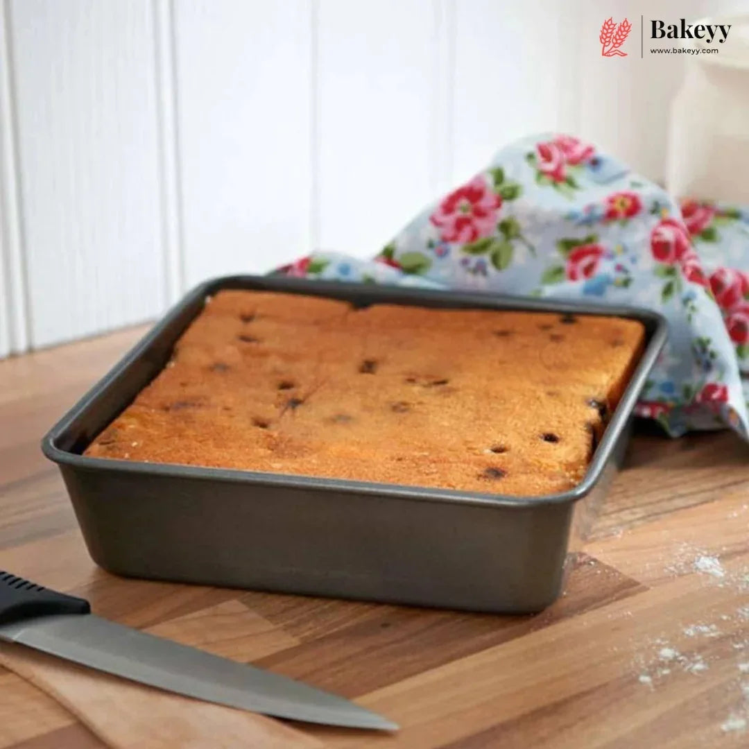 A rectangular dark gray baking pan containing a golden-brown rectangular cake, set on a wooden surface with a floral cloth and a knife nearby. The Bakey brand logo is visible in the top right corner.