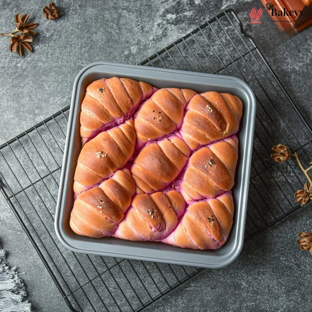 A square gray loaf pan filled with twisted, baked bread rolls topped with brown sugar and sprinkled with seeds, resting on a wire cooling rack over a gray stone surface with star anise and a small bottle in the corner.