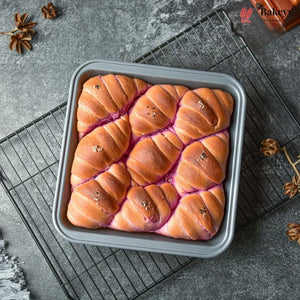 A square gray loaf pan filled with twisted, baked bread rolls topped with brown sugar and sprinkled with seeds, resting on a wire cooling rack over a gray stone surface with star anise and a small bottle in the corner.