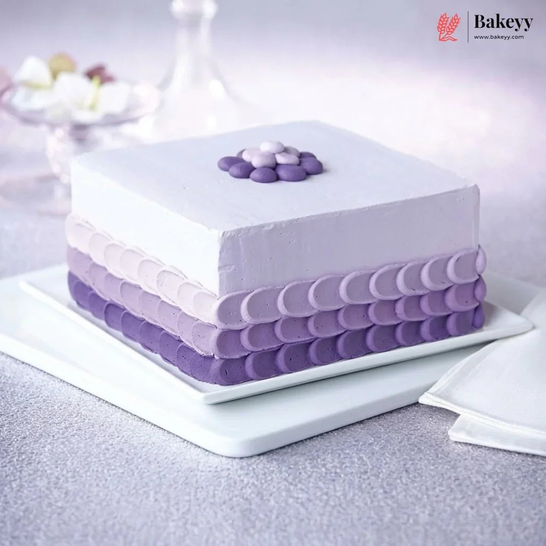 A square frosted cake in shades of purple and lavender, decorated with piped circular borders and a small flower on top, presented on a white plate against a soft, light purple background.
