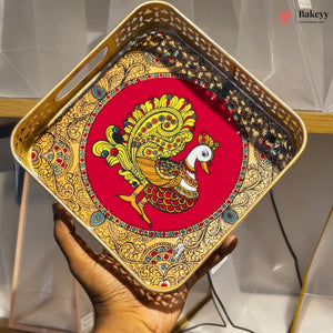 A square decorative tray with cutout handles, held in a hand. The tray features an ornate gold-toned border with red, green, and blue accents, surrounding a central red circle with a detailed gold, orange, and white peacock illustration.