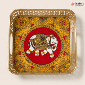 A square decorative tray with a gold-toned metal frame and side handles, featuring a hand-painted style elephant motif on a red circular background with ornate yellow floral patterns around the border. The Bakeyy brand logo is visible in the top right corner.