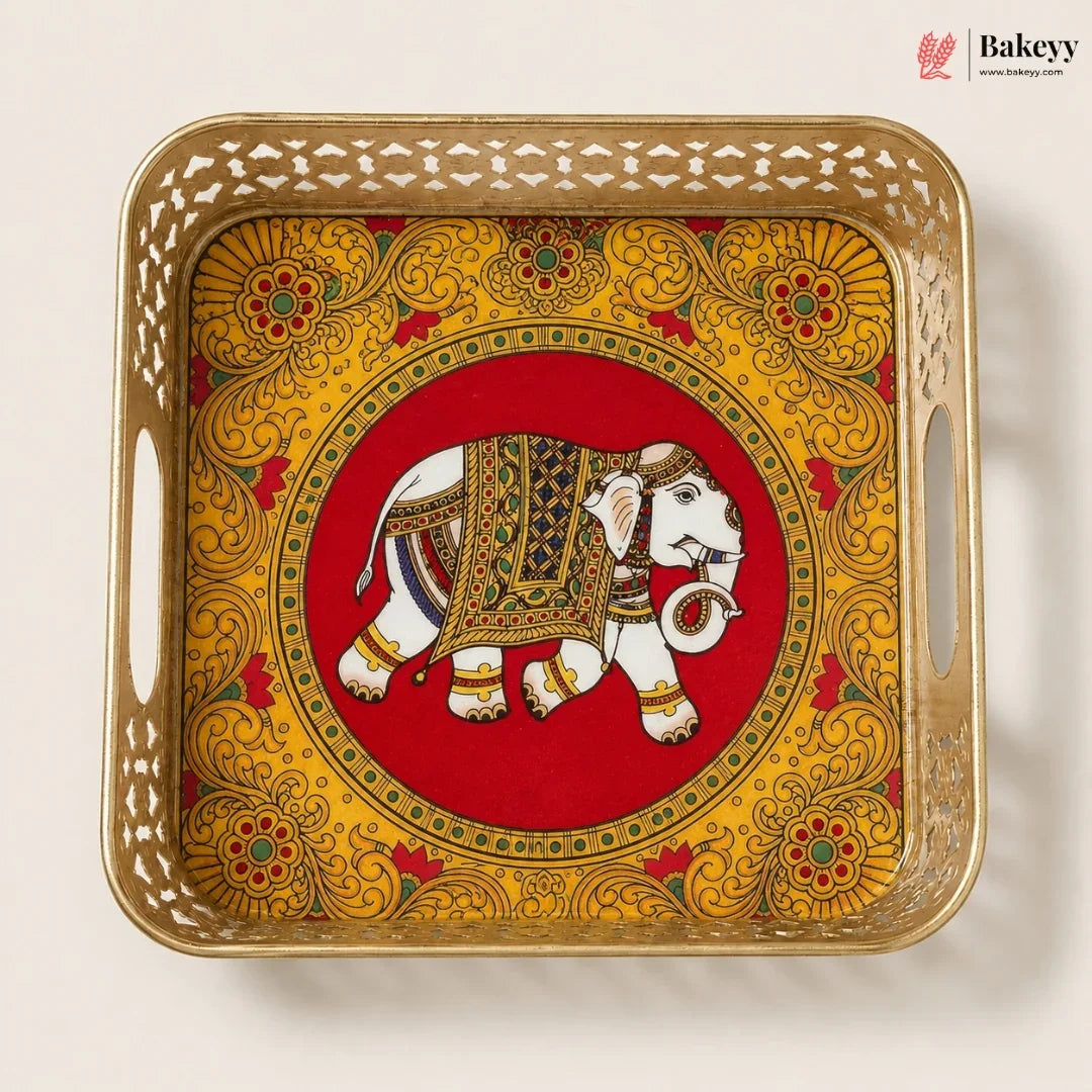 A square decorative tray with a gold-toned metal frame and side handles, featuring a hand-painted style elephant motif on a red circular background with ornate yellow floral patterns around the border. The Bakeyy brand logo is visible in the top right corner.