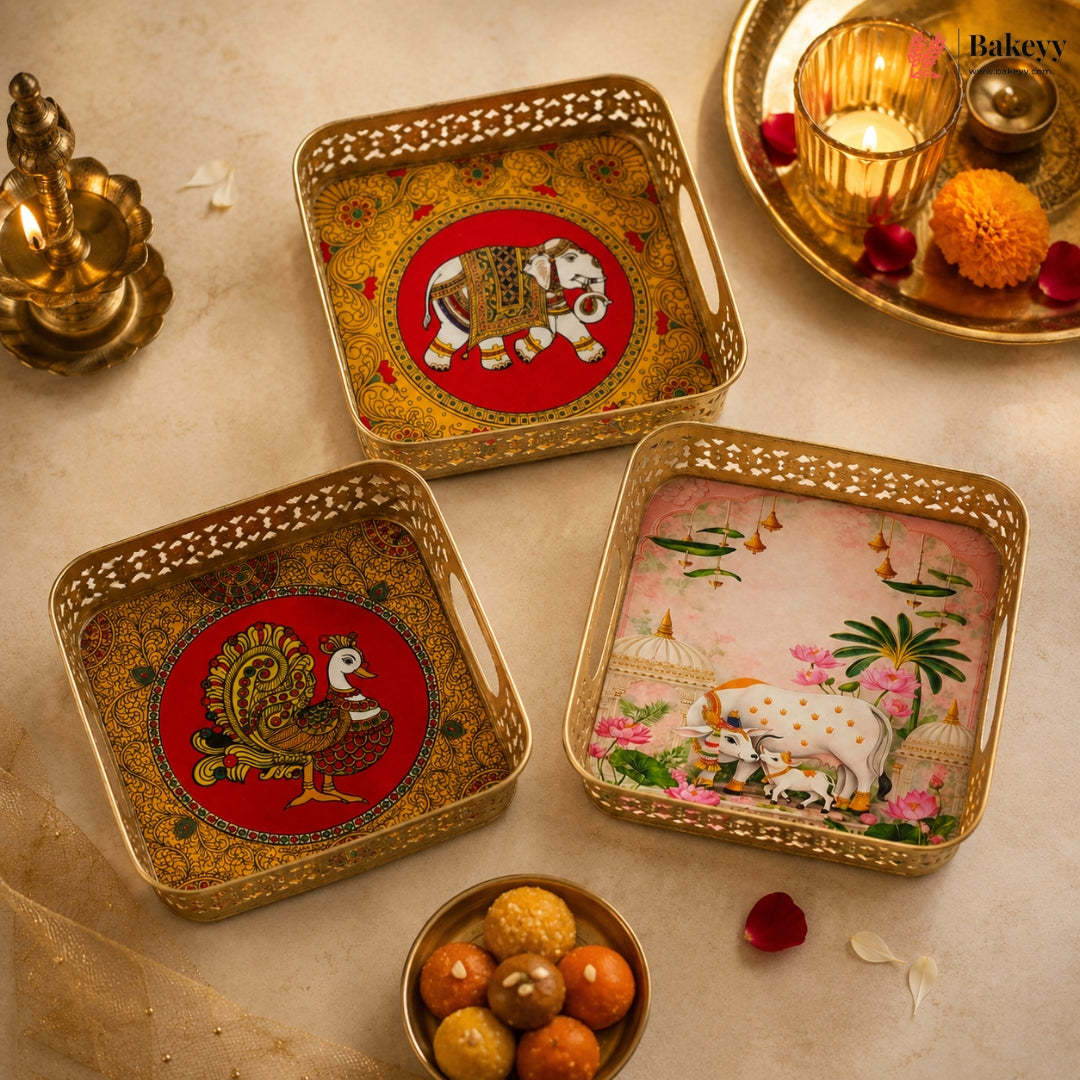 Three decorative square metal trays with ornate cutout edges, arranged on a beige surface with festive props. Each tray has a gold-toned border and a central painted motif: one red circle with an elephant, one red circle with a peacock, and one pink scene with cows and flowers.