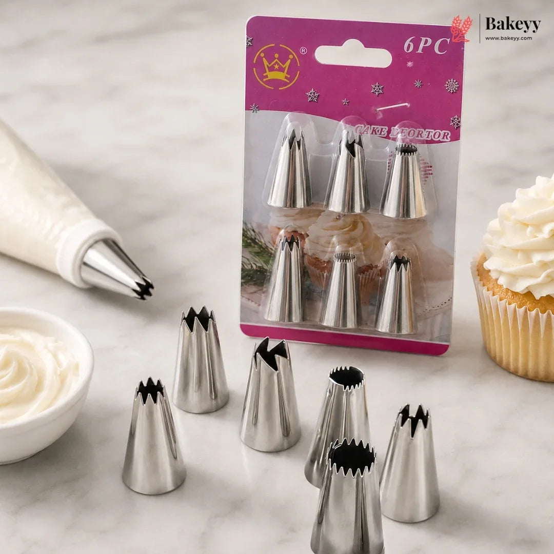 Six metal cupcake piping tips displayed on a marble surface, with one tip mounted on a piping bag and others shown both inside their pink retail blister pack and loose. A cupcake with white frosting and a small bowl of frosting are placed around them as styling props.