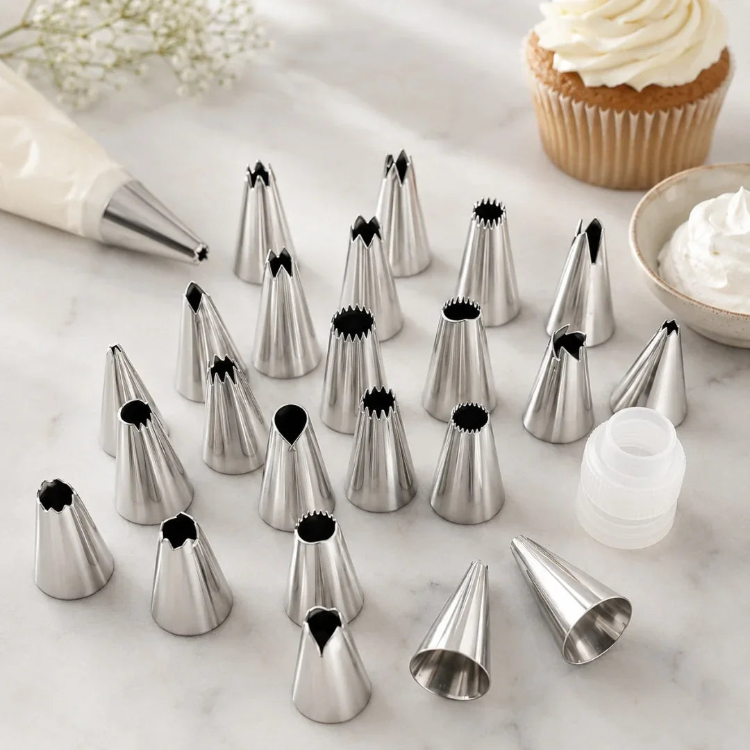 A set of stainless steel pastry piping tips arranged on a white marble surface, with a piping bag, a cupcake with white frosting, and a small bowl of frosting in the background.