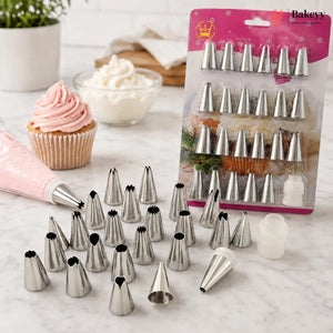 A set of metal piping tips displayed on a marble surface, with a pink piping bag and a packaged set of baking tips in the background. A cupcake with pink frosting and bowls of frosting are also visible.