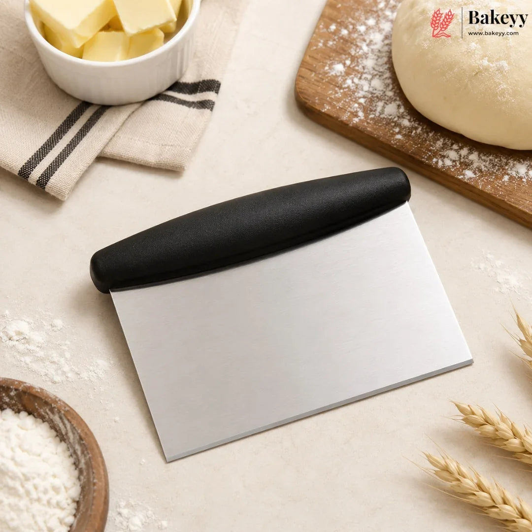 A stainless steel pastry scraper with a black handle, styled on a beige surface with baking ingredients including flour, butter, dough, and wheat stalks. The Bakey brand logo is visible in the top right corner.