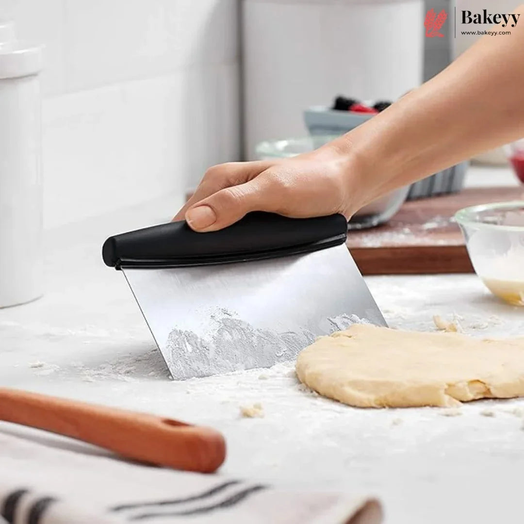 A hand using a metal pastry scraper with a black handle to remove dough from a floured surface in a kitchen setting.