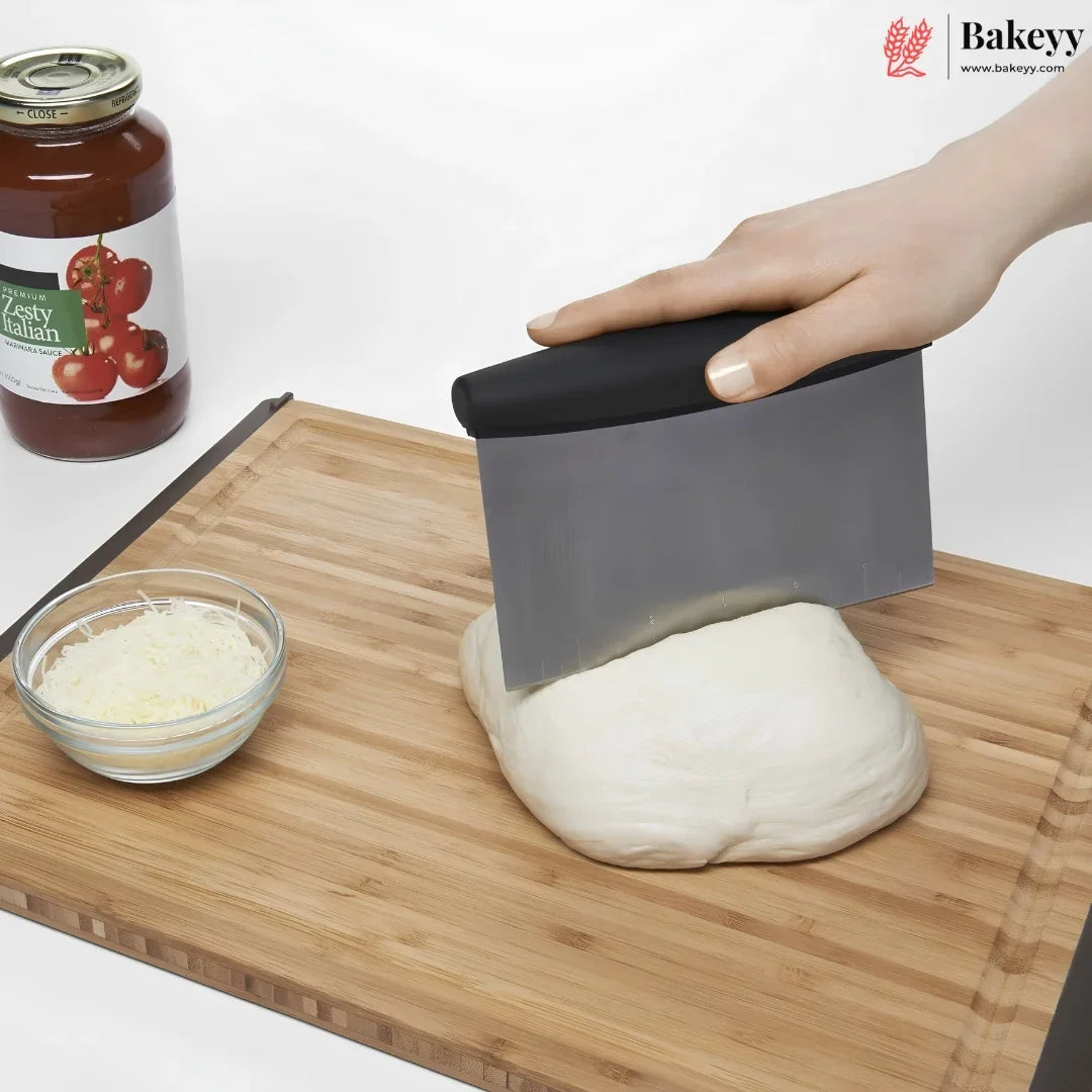 A hand uses a gray and black bench scraper to cut a ball of dough on a wooden cutting board, with a jar of tomato sauce and a small bowl of shredded cheese in the background. The Bakeyy logo is visible in the top right corner.