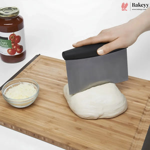 A hand uses a gray and black bench scraper to cut a ball of dough on a wooden cutting board, with a jar of tomato sauce and a small bowl of shredded cheese in the background. The Bakeyy logo is visible in the top right corner.