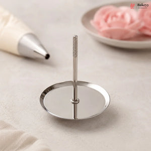 A stainless steel cake stand with a single upright stem and a round flat base, shown on a light neutral surface with a piping bag and pink frosting roses in the background.