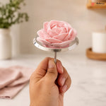 A hand holding a pink rose-shaped cake or pastry on a small round metal stand, with a softly blurred kitchen or bakery background and a visible Bakeyy logo in the top right corner.