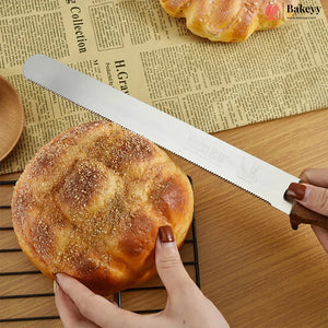 A serrated bread knife with a silver blade and dark handle being used to slice a round, golden-brown baked bread on a wooden table, with hands visible holding the bread and knife.