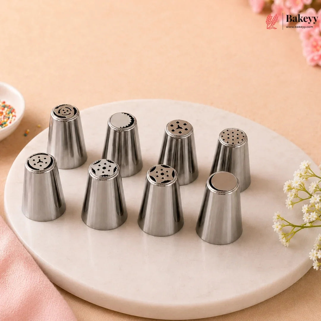 Eight stainless steel piping nozzles arranged on a round white marble tray against a warm beige background with soft pink floral accents. A Bakeyy logo appears in the top right corner.