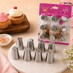 A 9-piece set of stainless steel piping nozzles displayed on a white marble plate, with their pink and gold retail blister packaging standing behind them. The scene is styled with cupcakes, a small bowl of sprinkles, and soft pink floral accents on a beige tabletop.