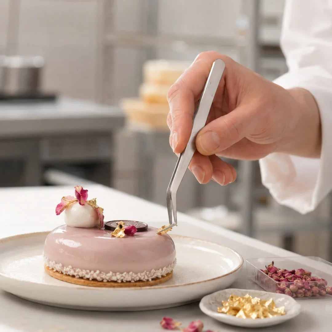 Person decorating a dessert with small flowers using tweezers in a kitchen setting.