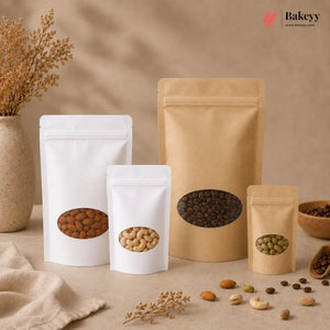 Four stand-up pouches of coffee beans in white and kraft paper packaging, arranged on a beige surface with dried flowers, beans, and a small bowl in a styled setting. The Bakeyy logo is visible in the top right corner.