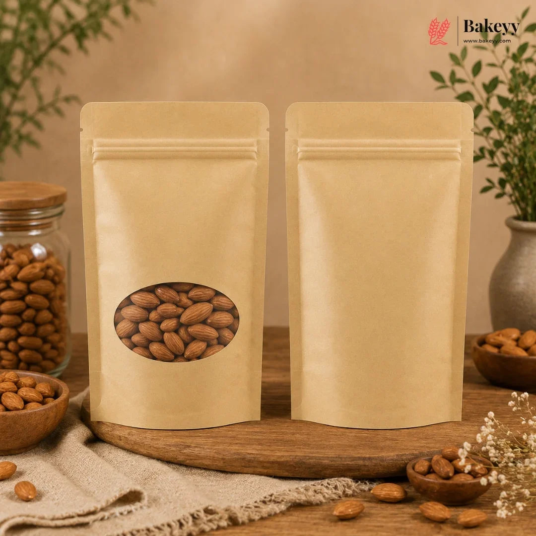 Two blank kraft paper stand-up pouches with one featuring an oval window showing almonds, displayed on a wooden board with almonds and a glass jar of almonds in a styled setting. The Bakey logo is visible in the top right corner.