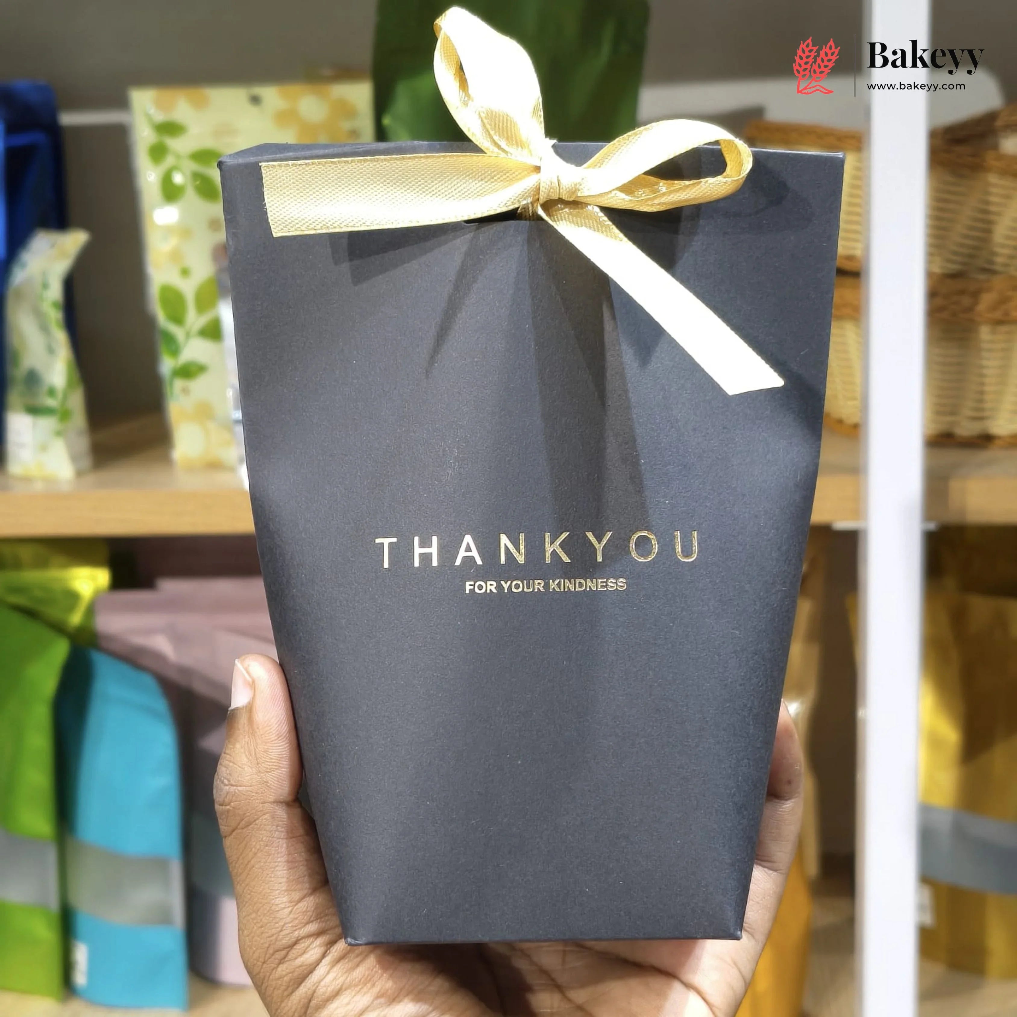 Blue gift box with a yellow ribbon held by a hand, with 'Bakeyy' branding in the background.