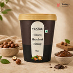 A 1kg tub of TENERO Choco Hazelnut Filling, featuring a dark brown label with a cream-colored center label and a gold lid. The tub is styled on a beige stone surface with hazelnuts, chocolate pieces, and a small bowl of the chocolate filling, under soft, warm lighting.