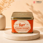 A square metal tin of Tiger Kesari Colour food coloring, displayed on a round beige pedestal. The tin has a metallic gold lid and a bright orange label with a white central badge.