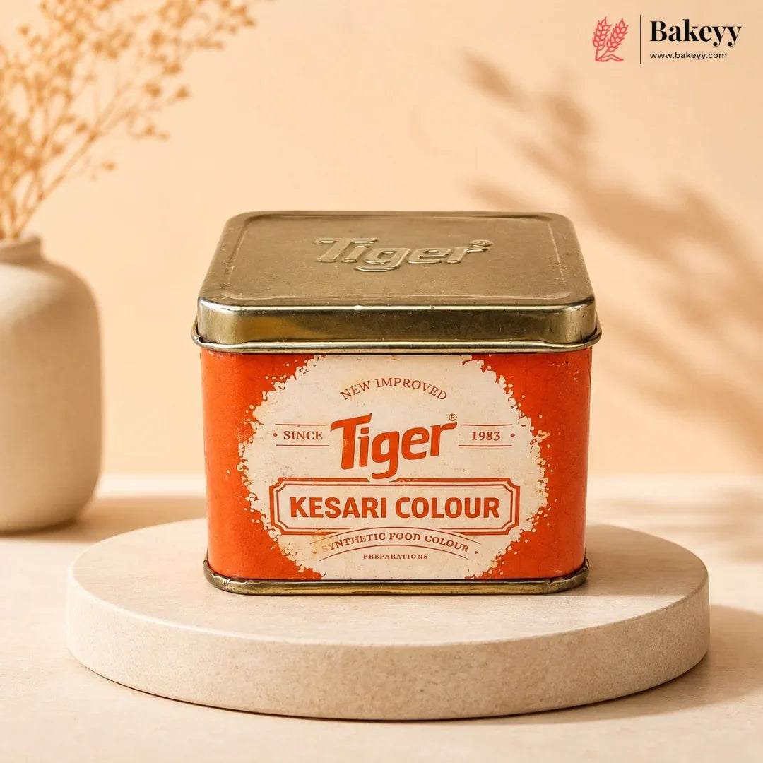 A square metal tin of Tiger Kesari Colour food coloring, displayed on a round beige pedestal. The tin has a metallic gold lid and a bright orange label with a white central badge.