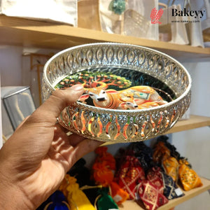A hand holds a round silver-toned metal tray with ornate cutout patterns and a colorful printed illustration of a deity on the bottom. The background shows wooden shelves with various items in a shop setting, and a 'Bakeyy' watermark is visible in the top right corner.
