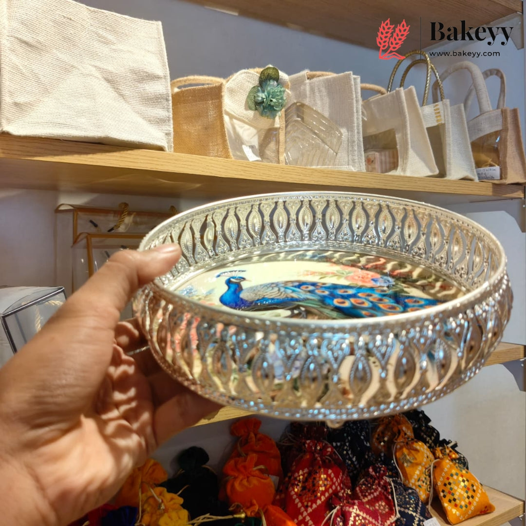 A hand holds a round silver-toned decorative tray with an intricate cut-out floral border and a colorful peacock illustration on the inner surface. The background shows shelves with fabric bags and other items in a shop setting, with a Bakeyy logo watermark in the top right corner.