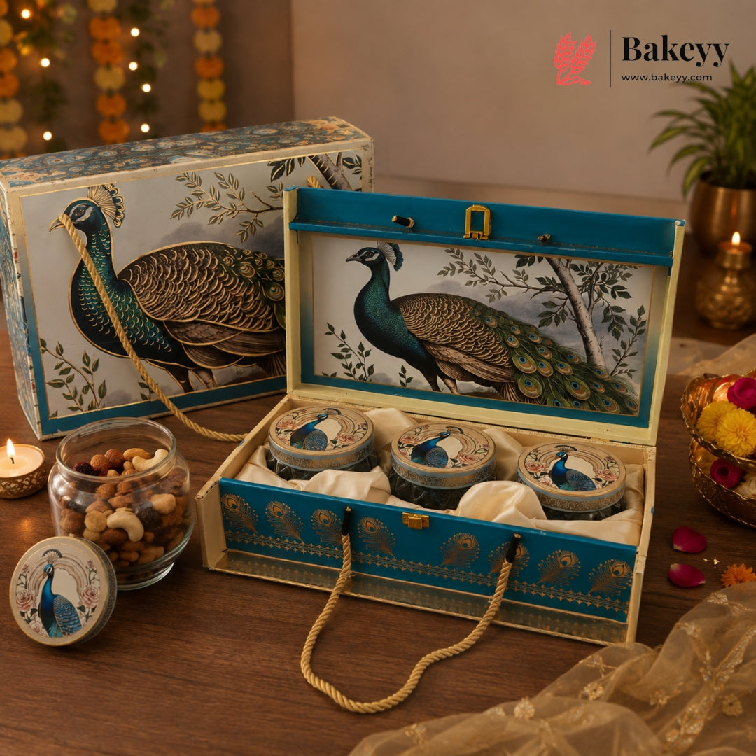 A decorative gift set featuring a large blue and cream peacock-themed box with three matching round tins inside, shown open on a wooden table. A smaller matching box sits behind it, and a small branded Bakeyy logo appears in the top right corner.