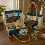 A decorative gift set featuring a blue and cream peacock-themed box with rope handles, an open matching box with a round tin and small jar inside, and a matching tote bag on a wooden table. The set includes printed peacock artwork, cream fabric lining, and glass containers.