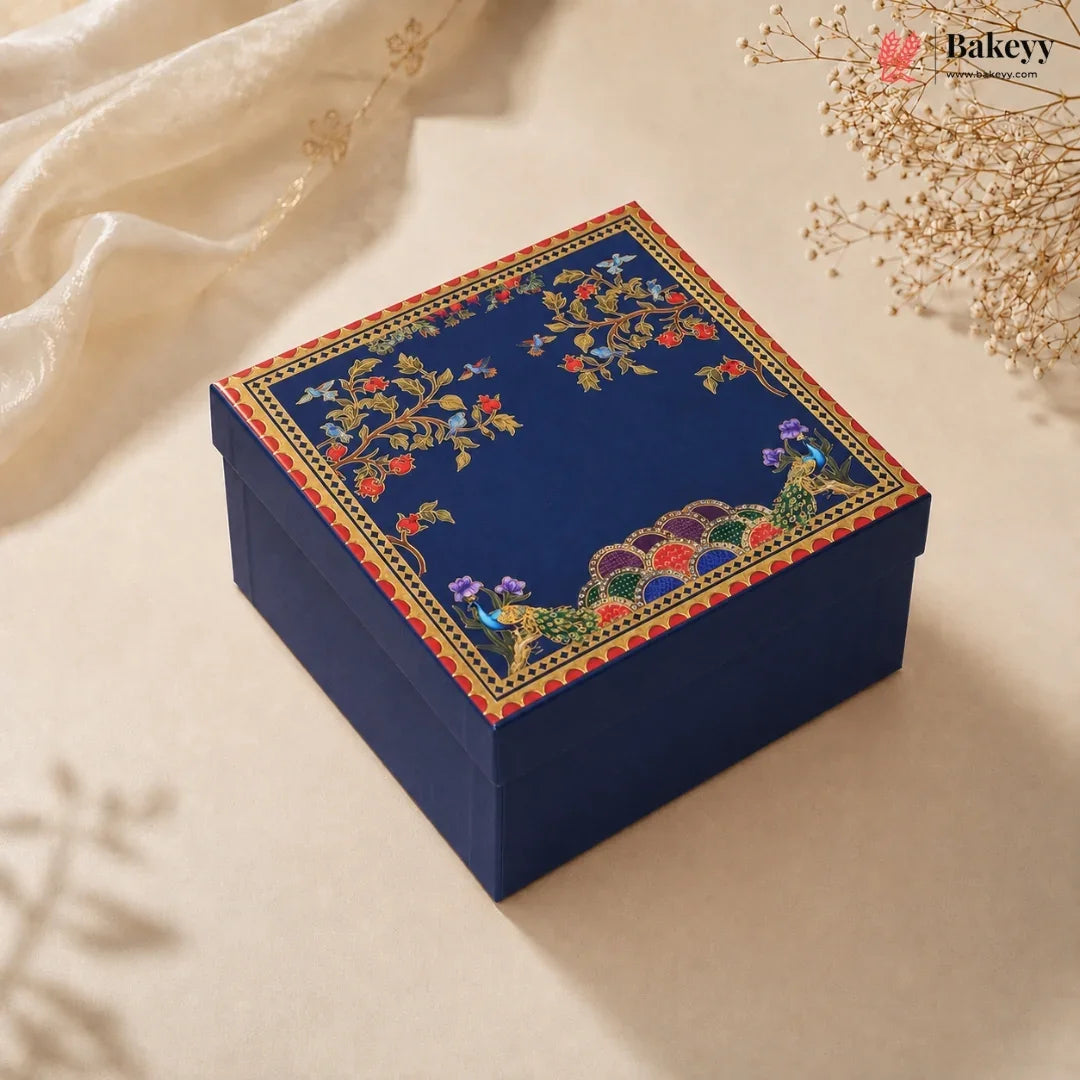 A square navy blue gift box with an ornate floral and peacock pattern on the lid, styled on a beige surface with soft cream fabric and dried flowers.