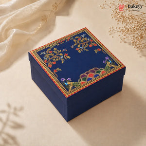 A square navy blue gift box with an ornate floral and peacock pattern on the lid, styled on a beige surface with soft cream fabric and dried flowers.