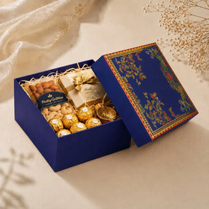 A luxury chocolate and nut gift box set in deep blue with gold foil Ferrero Rocher chocolates, cashews, almonds, and a small wrapped present inside, shown on a beige surface with decorative dried flowers and draped fabric.