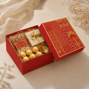 A luxury gift box set containing a red patterned box with its lid off, revealing a gift basket with almonds, a wrapped cream box, and Ferrero Rocher chocolates, styled on a beige surface with decorative dried flowers.