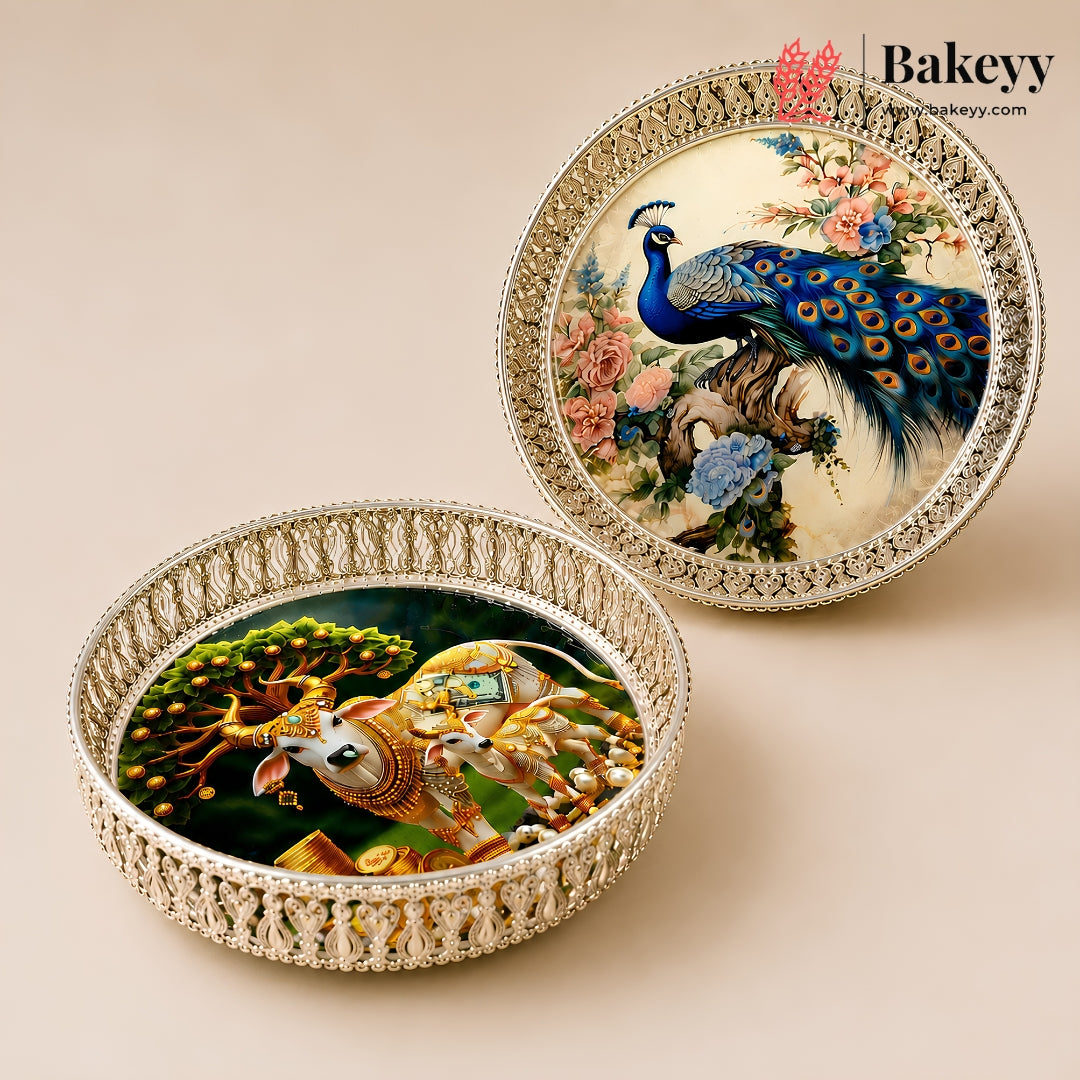 Two round decorative trays with ornate silver-toned borders, one featuring a peacock and floral scene, and the other featuring a golden elephant and tree scene, displayed on a beige background. The Bakeyy logo is visible in the top right.