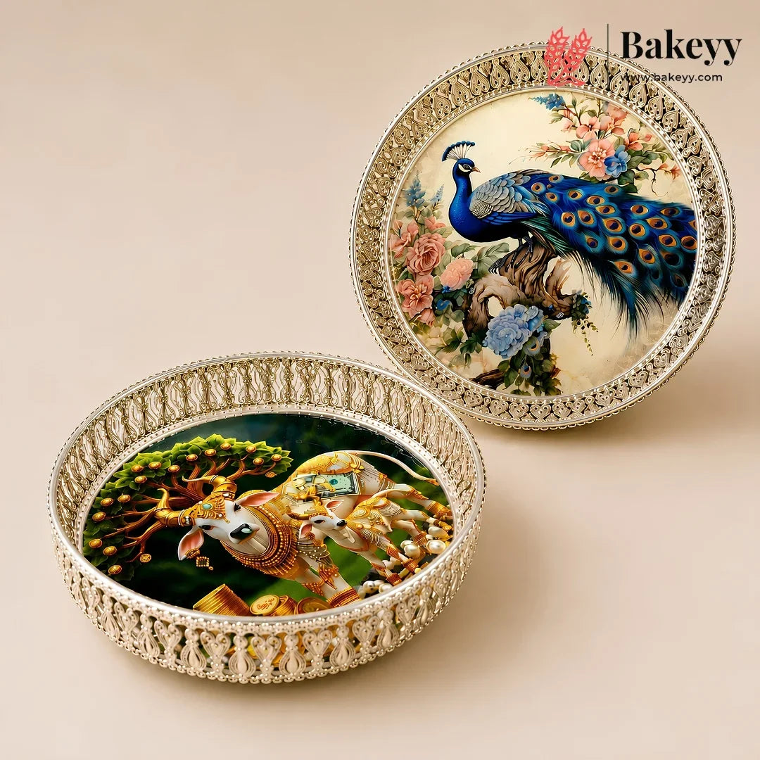 Two round decorative trays with ornate silver-toned borders, one featuring a peacock and floral scene, and the other featuring a golden elephant and tree scene, displayed on a beige background. The Bakeyy logo is visible in the top right.