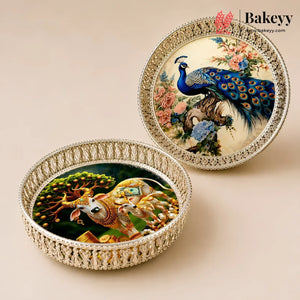 Two round decorative trays with ornate silver-toned borders, one featuring a peacock and floral scene, and the other featuring a golden elephant and tree scene, displayed on a beige background. The Bakeyy logo is visible in the top right.