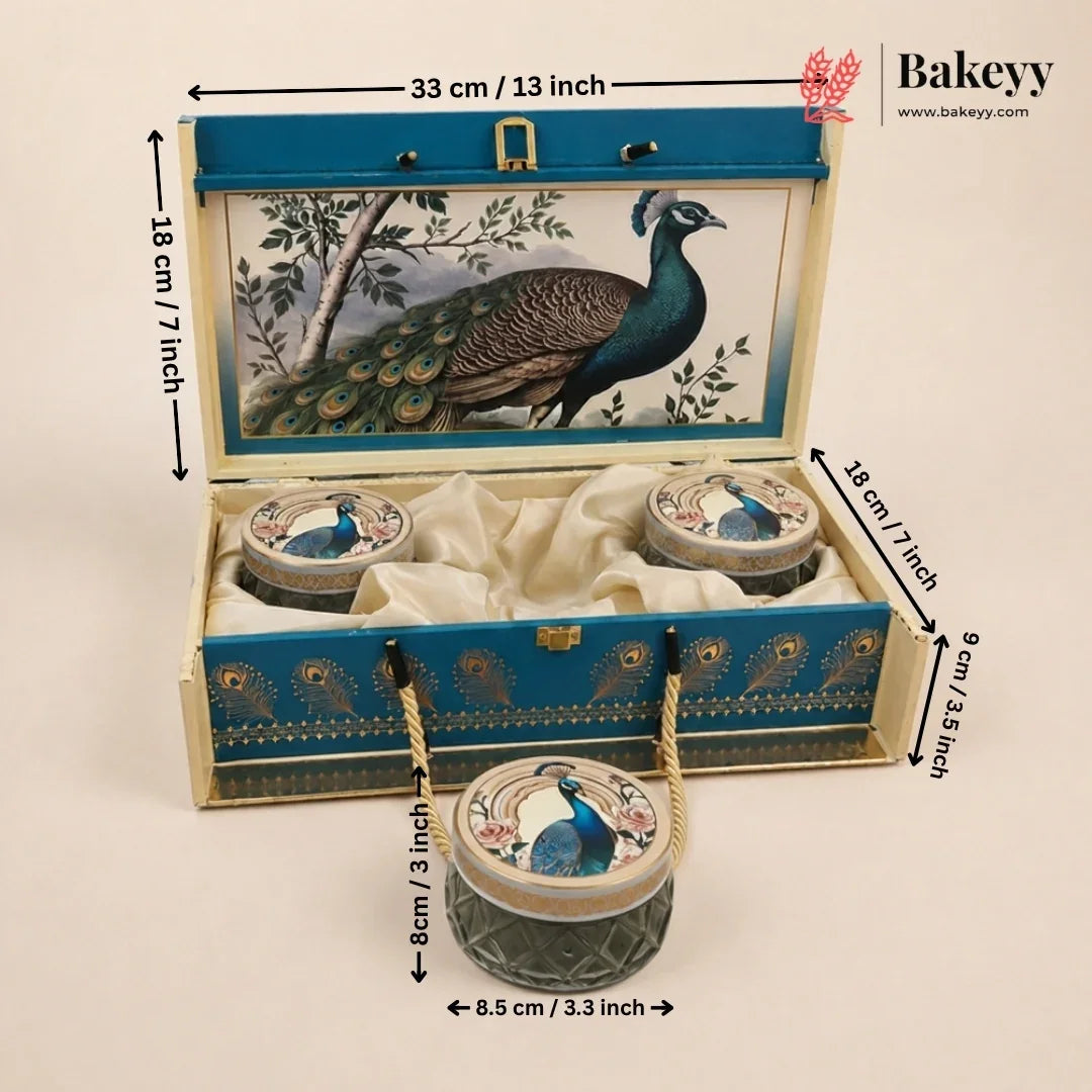 An open decorative Bakeyy gift box for tea or biscuits, shown with dimension callouts. The box is cream-colored wood with a blue lid panel featuring a peacock illustration, velvet lining, and three small round containers attached by rope handles.